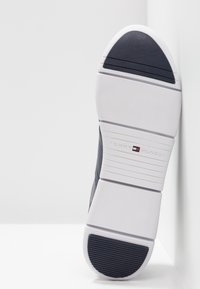 Semelle de basket blanche avec zones d'adhérence noires au talon et à l'avant, portant le logo Tommy Hilfiger et le nom de la marque embossés au centre.