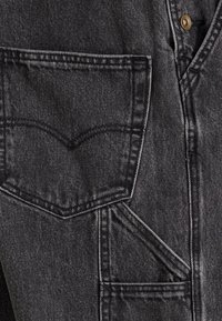 Κοντινή λήψη μαύρου τζιν υφάσματος που δείχνει λεπτομέρειες ραφής, πίσω τσέπη και μεταλλικό κουμπί στη μέση.