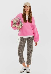 Przesadnie duży różowy sweter z dzianiny, biała koszula pod spodem, spodenki w panterkę do kostek, trzymając bukiet małych kwiatów i nosząc beżową czapkę.