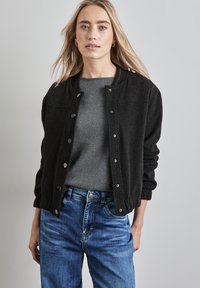 Giubbotto bomber nero con chiusura a bottoni a pressione, colletto a coste e polsini elasticizzati, abbinato a un maglione grigio e jeans in denim blu.