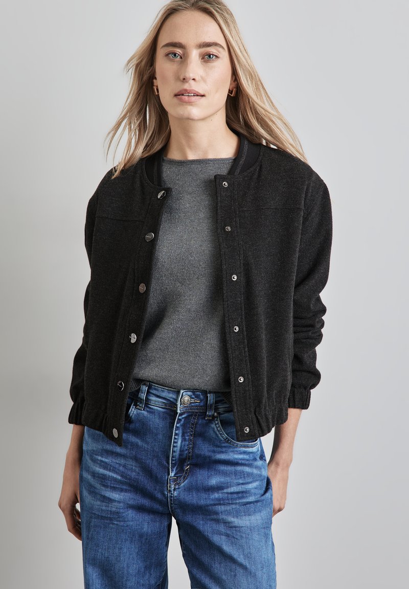 Giubbotto bomber nero con chiusura a bottoni a pressione, colletto a coste e polsini elasticizzati, abbinato a un maglione grigio e jeans in denim blu.