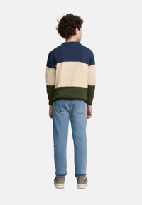 Jersey con bloques de color en azul marino, crema y verde oliva; niño que lleva vaqueros de mezclilla azul claro, zapatos grises y beige, y con el pelo rizado visible.