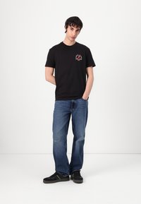 Black cotton t-shirt with a round neckline featuring a red and white graphic design on the chest. Paired with light blue denim jeans.