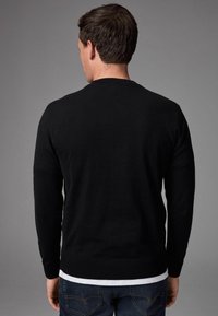 Zwarte gebreide trui met lange mouwen en een ronde hals, met ribgebreide manchetten en zoom. Draagt over een witte blouse, achteraanzicht weergegeven.