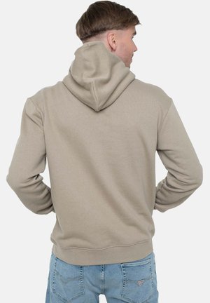 Uomo con felpa con cappuccio beige e jeans azzurri, di spalle con le mani sui fianchi contro uno sfondo semplice.