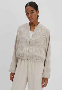 Beige Bomberjacke mit strukturiertem, gefälteltem Design, ausgestattet mit einem frontalen Reißverschluss, gerippten Bündchen und einem lockeren Schnitt. Leichtes Material.