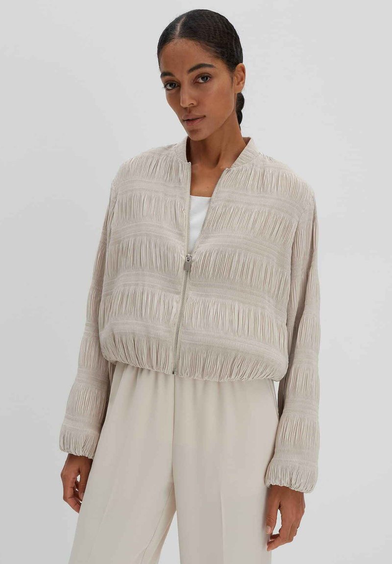 Beige Bomberjacke mit strukturiertem, gefälteltem Design, ausgestattet mit einem frontalen Reißverschluss, gerippten Bündchen und einem lockeren Schnitt. Leichtes Material.