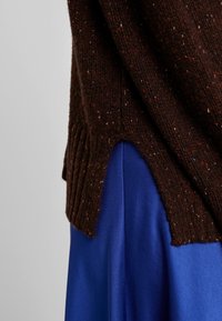 Brun stickad tröja med ribbad textur och prickar, med en slits på sidan. Matchas med ett slätt, klarrött tyg nedanför.