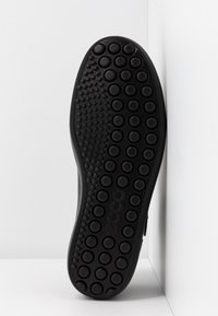 Semelle de chaussure noire avec motif circulaire de la bande de roulement et points texturés variés, positionnée verticalement contre un fond blanc en coin.