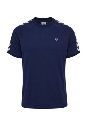 Ung mand iført en marineblå t-shirt med hvide chevronstriber på ærmerne og en matchende to-farvet kasket, kigger nedad med hånden let løftende på t-shirten.