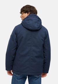 Námořnická modrá bunda s kapucí a hladkým povrchem, která má uvolněný střih a nenápadné švy. Na zadní straně je kapuce a jednoduchý design.