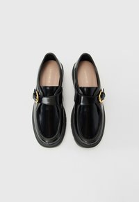 Pair of black glossy leather loafers with gold buckles and thick rubber soles, beige insoles branded "COCCINELLE," on a white background.