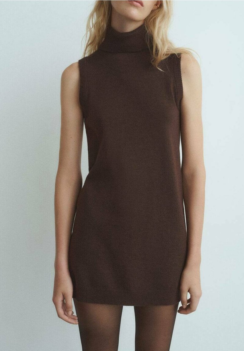 Robe mini marron en maille sans manches à col roulé portée par une personne debout à la peau claire et aux cheveux blonds sur un fond uni.