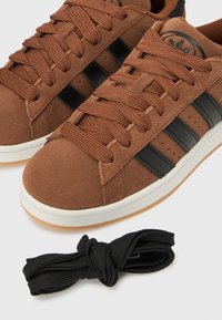 Hnedé semišové tenisky s čiernymi pruhmi, textúrovanými šnúrkami a bielou gumovou podrážkou. Súčasťou balenia je zviazaná čierna šnúrka.