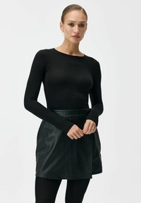 Czarna bluzka z długim rękawem i okrągłym dekoltem zestawiona z ciemnozieloną skórzaną mini spódnicą. Spódnica ma owijany fason i gładką fakturę.