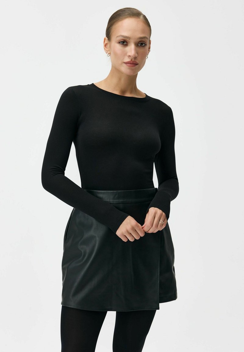 Czarna bluzka z długim rękawem i okrągłym dekoltem zestawiona z ciemnozieloną skórzaną mini spódnicą. Spódnica ma owijany fason i gładką fakturę.