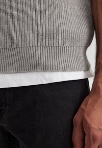 Grauer gerippter Strickpullover mit einem enganliegenden Saum, der über einem weißen Hemd getragen wird, kombiniert mit dunklen Jeans. Fokus auf Texturen und dem geschichteten Design.
