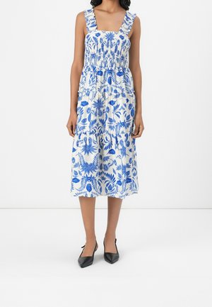 Femme portant une robe midi blanche avec imprimé floral bleu, bretelles à volants et corsage froncé, associée à des chaussures slingback noires à bout pointu.