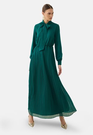Donna che indossa un lungo vestito plissettato color verde acqua con scollo a fiocco e scarpe dorate con punta, in piedi davanti a uno sfondo chiaro uniforme.