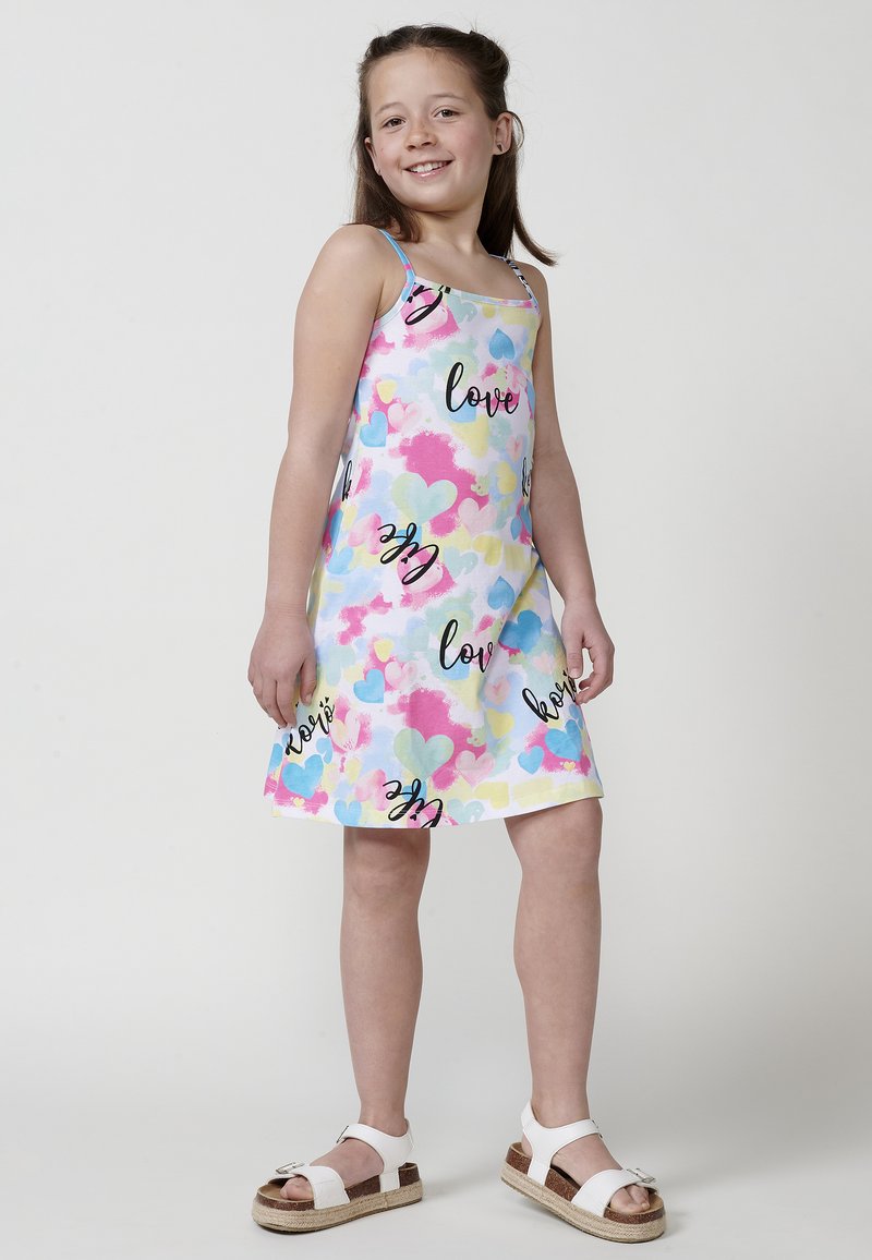 Weißes Kleid mit einem bunten Herzmuster in Pink, Blau und Gelb, versehen mit schwarzem "Love"-Text. Kombiniert mit weißen Sandalen.