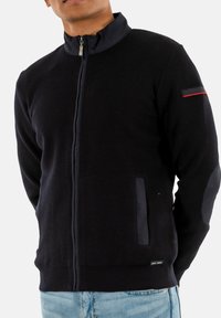 Chaqueta negra de punto con cremallera y cuello alto, que presenta un acento rojo en el hombro izquierdo y bolsillos laterales con acentos negros.