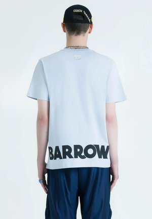 Persona que lleva una camiseta azul clara con el texto "BARROW" en la parte baja de la espalda, pantalones azul marino y una gorra negra con la etiqueta "COACH", mirando hacia otro lado.
