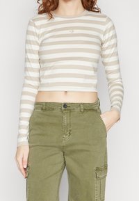 Long-sleeve, cropped top in beige with white horizontal stripes. Features a round neckline and a small logo on the chest. Soft fabric.