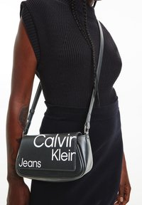 Černá kožená kabelka přes rameno s prohnutým tvarem a bílým logem „Calvin Klein Jeans“. Nastavitelný popruh a hladká textura.