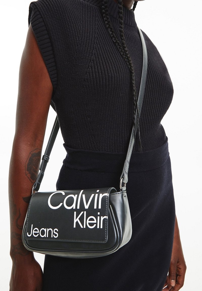 Černá kožená kabelka přes rameno s prohnutým tvarem a bílým logem „Calvin Klein Jeans“. Nastavitelný popruh a hladká textura.