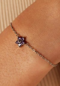 Armband met een zilveren ketting in bloemenontwerp, versierd met paarse edelstenen in een clusterpatroon. Gestructureerde huid zichtbaar.