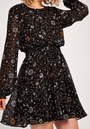 Vestido negro de manga larga con tela transparente, con patrones celestiales en blanco y dorado de estrellas, soles, lunas y constelaciones, usado por una persona.