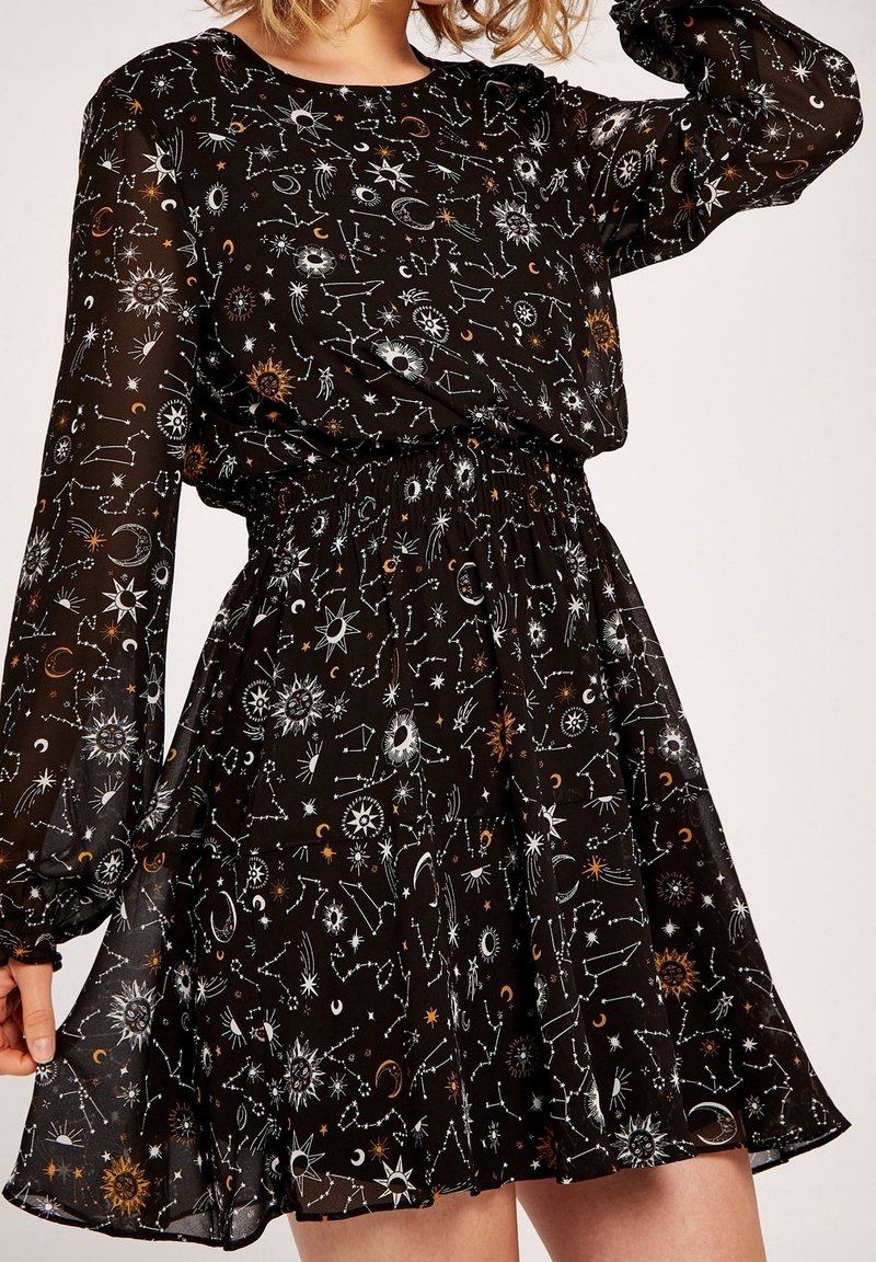 Robe noire à manches longues en tissu transparent, avec des motifs célestes blancs et dorés d'étoiles, soleils, lunes et constellations, portée par une personne.