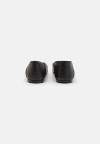 Zapatillas planas de cuero negro con una textura suave, punta redondeada y un sutil logotipo en el tacón. Diseño minimalista, sin herrajes visibles.