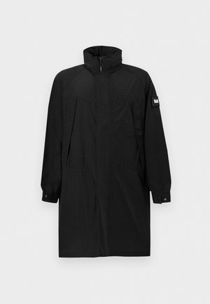 Manteau long noir avec col montant, fermeture éclair frontale, poches latérales, poignets à boutons et un écusson sur le haut de la manche gauche sur fond blanc.