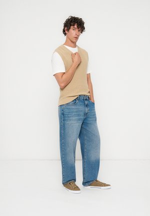 Hombre con cabello rizado que lleva un suéter sin mangas beige, camiseta blanca, vaqueros azules sueltos y zapatos deportivos marrones, de pie contra un fondo blanco.