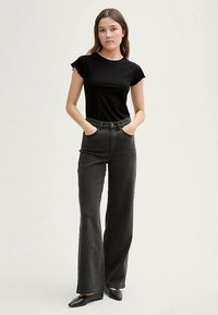 Czarny t-shirt z krótkim rękawem w połączeniu z wysokim stanem, rozszerzanymi ciemnoszarymi dżinsami. Modelka pozuję z rękami w kieszeniach, ma na nogach czarne buty.