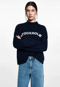 Mörkblå stickad tröja med texten "STOCKHOLM" i vitt, rund halsringning, avslappnad passform, kombinerad med ljusblå denimjeans med lapplådor.