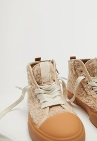 Béžové vysoké tenisky s chlupatou texturou, šněrováním a hnědými gumovými podrážkami. Obsahují boční zip a detaily s logem.