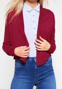 Burgundy cardigan made of soft knit, open front design, long sleeves. Worn over a light blue collared shirt with button details and denim jeans.