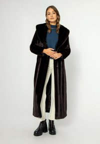 Dlouhý, tmavě hnědý kožich z umělé kožešiny s šálovým límcem, s rovným střihem, nošený přes modrý top a bílé kalhoty, doplněný černými platformovými botami.