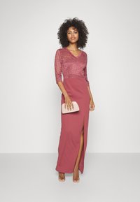 Vestido maxi em burgundy com corpete de renda e mangas três quartos, apresentando uma fenda lateral. A modelo segura uma clutch rosa claro.