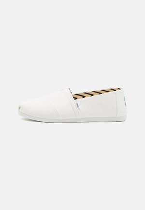 Zapatillas de lona blancas sin cordones con punta redonda, que cuentan con una suela de goma texturizada y un forro de tela a rayas en tonos neutros.