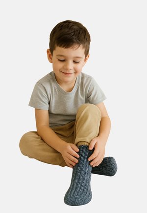 Junger Junge sitzt im Schneidersitz, lächelt sanft, trägt ein graues T-Shirt, beige Hosen und zieht dunkelgraue gestrickte Socken an.