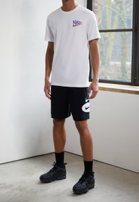 T-shirt en coton blanc avec un logo multicolore Nike sur la poitrine, associé à un short noir présentant un grand swoosh Nike blanc, et des baskets noires.