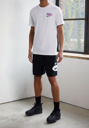 Weißes Baumwoll-T-Shirt mit mehrfarbigem Nike-Logo auf der Brust, kombiniert mit schwarzen Shorts, die einen großen weißen Nike-Swoosh zeigen, und schwarzen Turnschuhen.