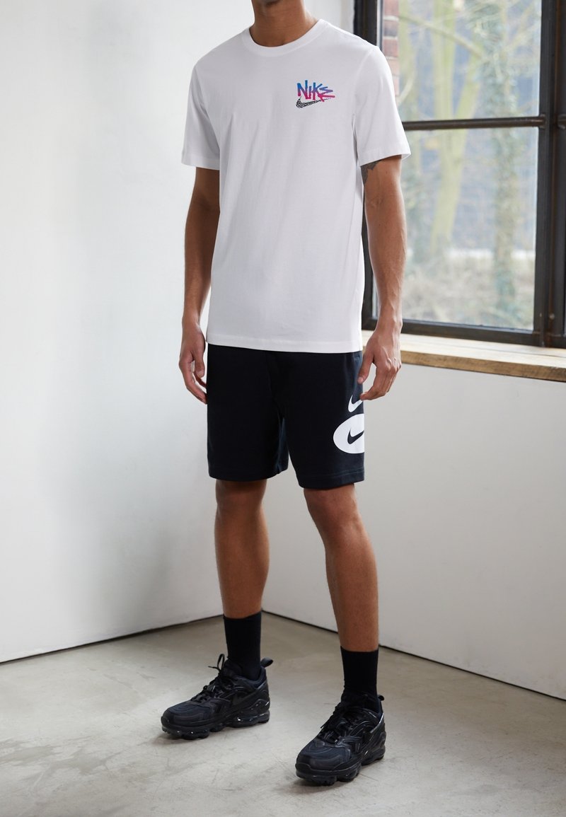 T-shirt en coton blanc avec un logo multicolore Nike sur la poitrine, associé à un short noir présentant un grand swoosh Nike blanc, et des baskets noires.