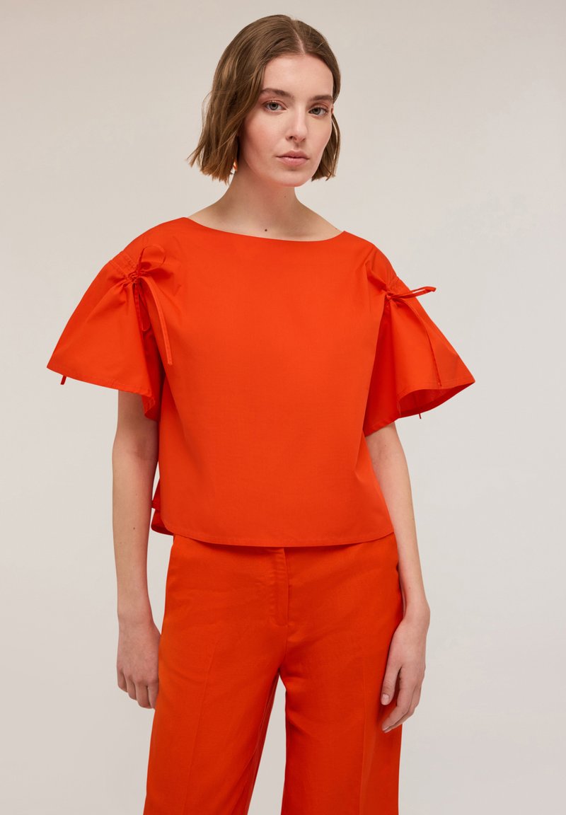 Top arancione a maniche corte con dettagli legati sulle maniche, caratterizzato da un design quadrato e un collo a barca, realizzato in tessuto morbido.