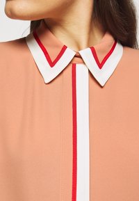 Blusa color pesca con colletto bianco bordato di rosso e striscia rossa verticale al centro. Tessuto morbido e liscio.