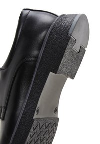 Zapato de cuero negro con una suela de goma texturizada, que presenta un tacón curvado y costuras visibles a lo largo de la parte superior elegante.