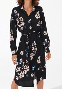 Černé květinové šaty s dlouhými rukávy, límečkem a páskem v oblasti pasu. Květinový vzor v růžových, bílých a modrých odstínech s knoflíkovými detaily.